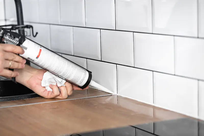 Cartridge of white silicone sealant being applied between white tiles and a wooden kitchen countertop