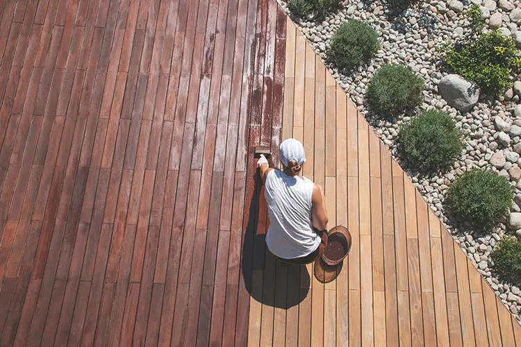 Overhead view of garden decking with person applying wood stain with a brush.