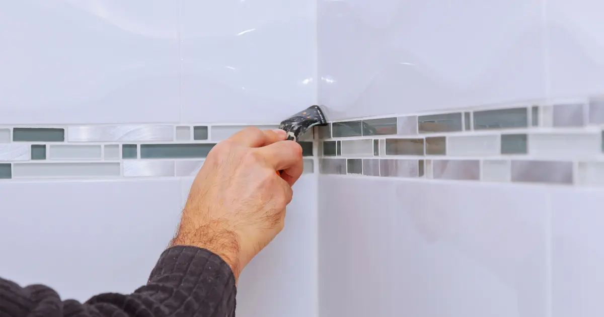 Sealing The Seam Between Ceramic Tiles A close-up of a person's hand using a sealant tool to apply white sealant to the corner of tiled walls with a decorative mosaic border.